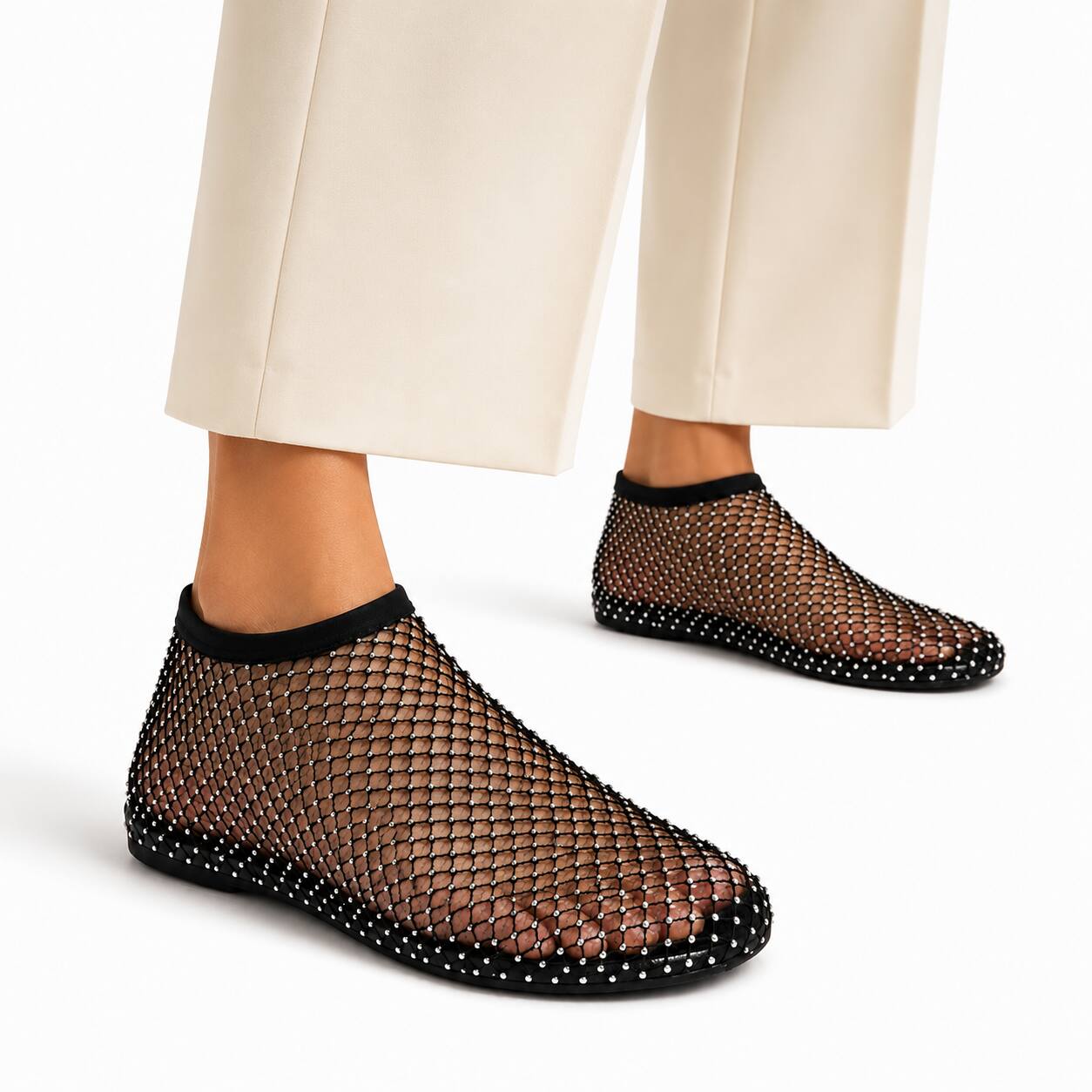 A person wearing black mesh slip-on shoes with white stud detailing around the edges and black trim, paired with cream-colored trousers, against a clean white background.
