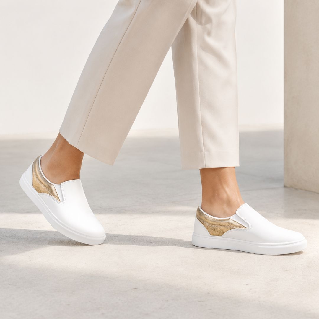 White slip-on sneakers with metallic gold wing-like accents on the sides, shown on a model wearing beige trousers in a minimalist concrete setting.