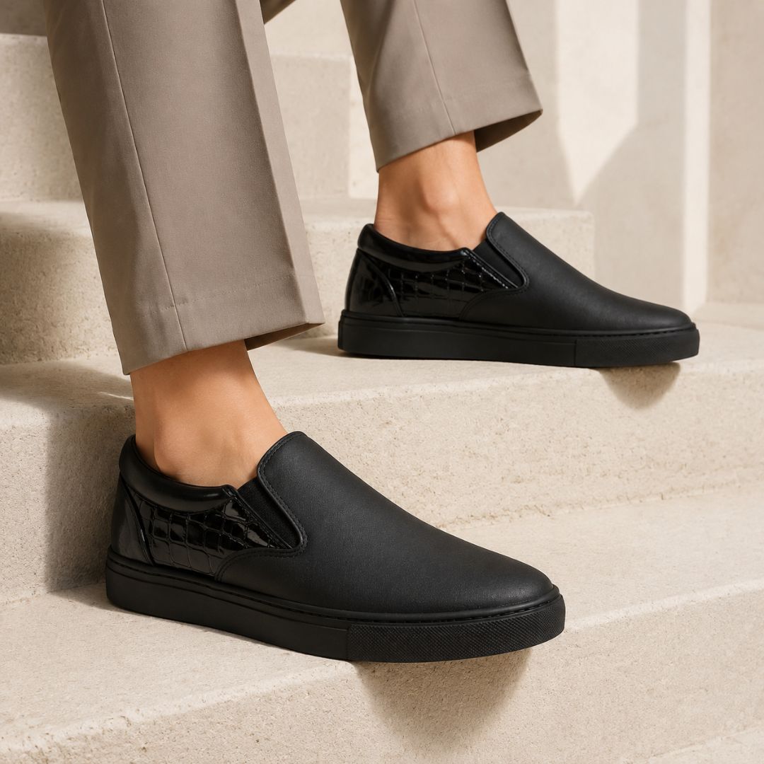 A person wearing black slip-on sneakers with a glossy crocodile-embossed heel detail, paired with beige trousers, standing on light beige stone steps.