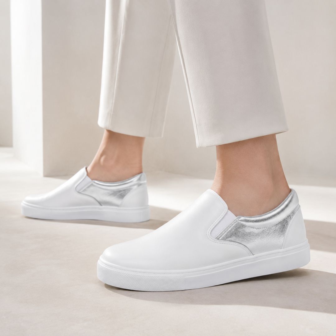 White slip-on sneakers with metallic silver side panels, shown on a model wearing off-white trousers in a bright, minimalist indoor setting.