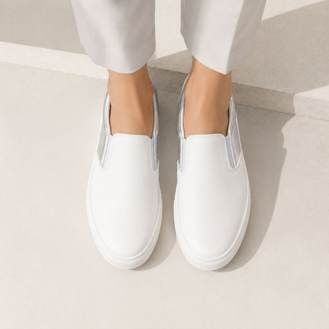 White slip-on sneakers worn with light gray trousers, shown from a top-down angle on a light concrete surface.