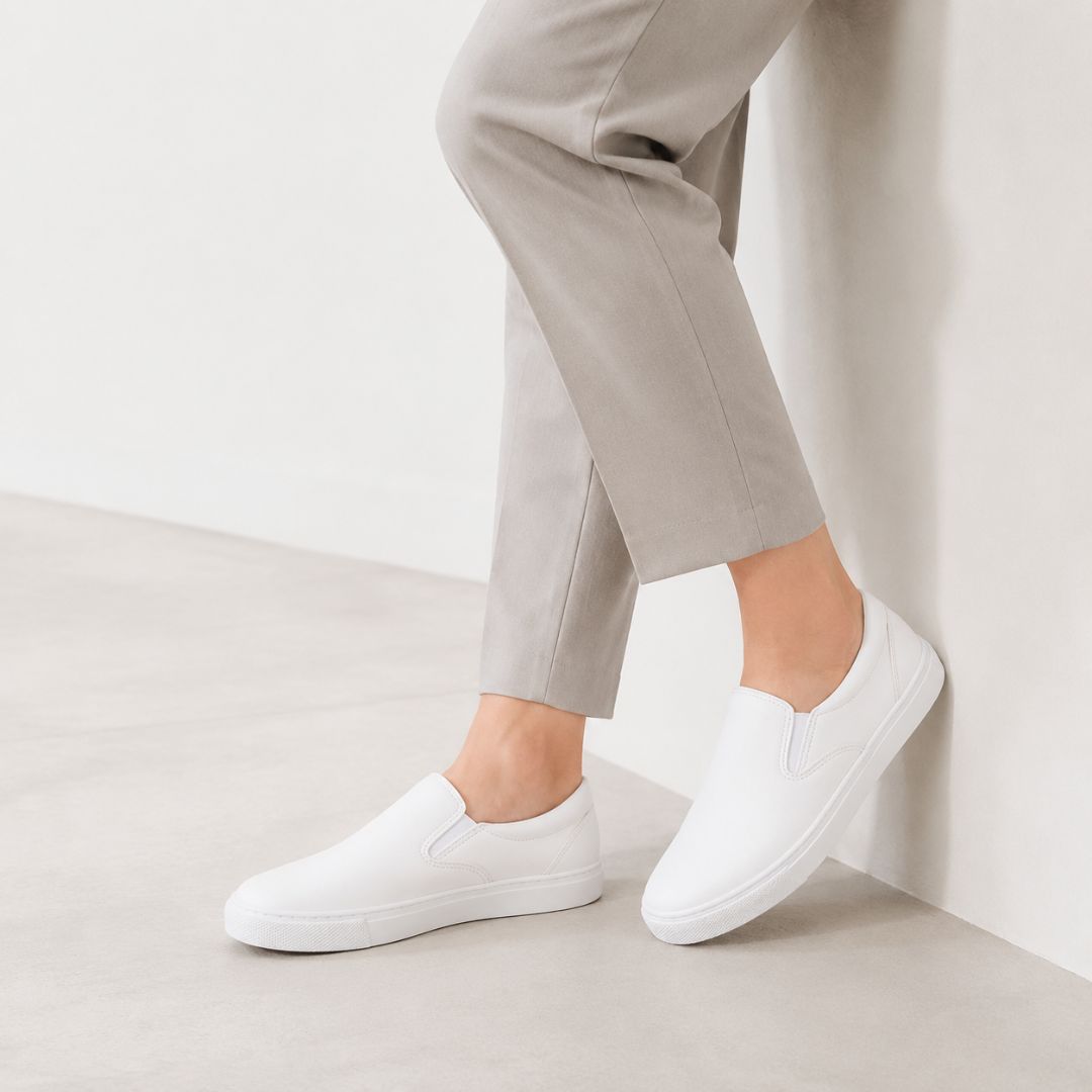 White slip-on sneakers worn with light beige trousers, shown on a model leaning against a white wall in a minimalist indoor setting.